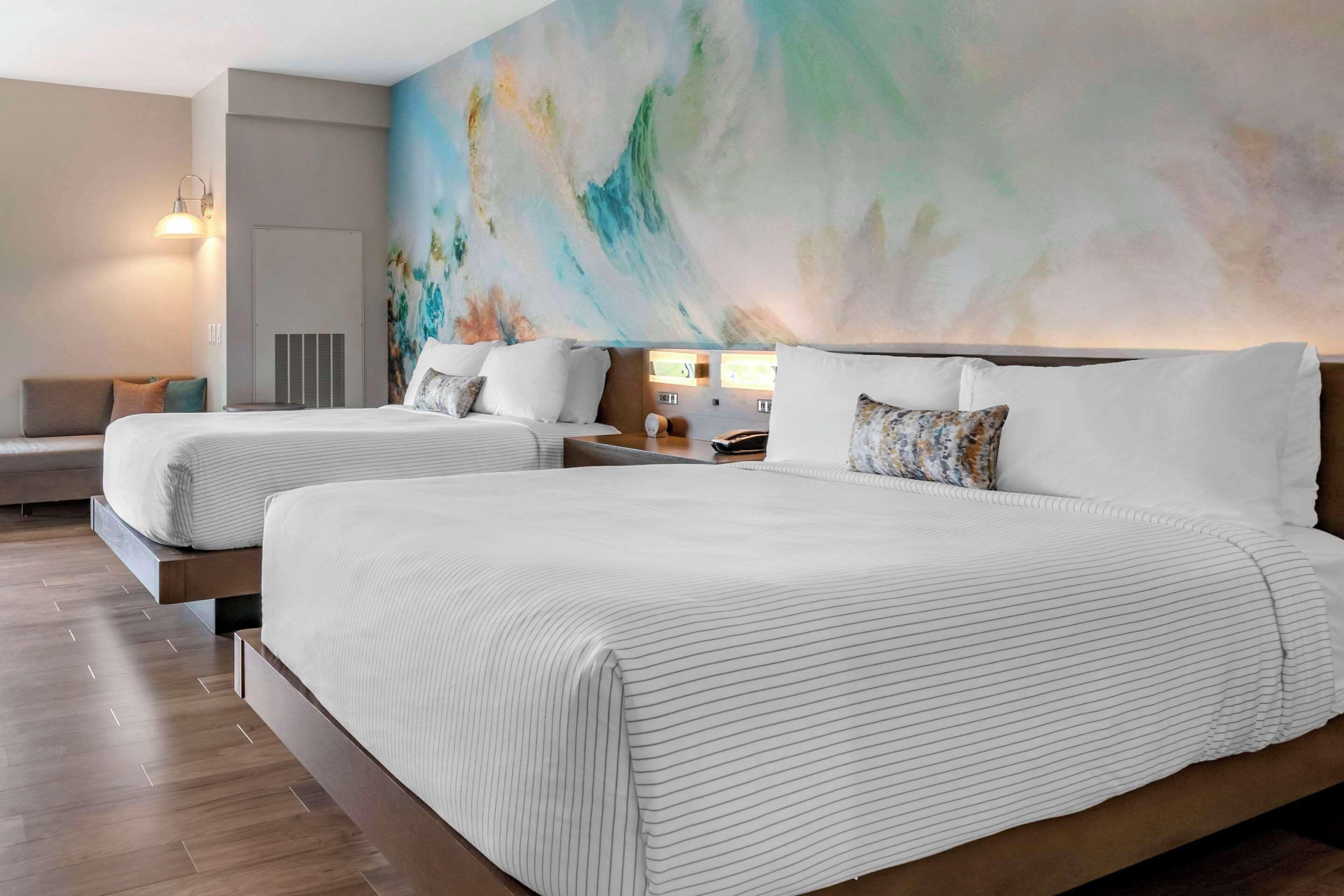 Habitación de hotel con dos camas, un gran mural abstracto en la pared y una zona de estar con sofá y mesa.