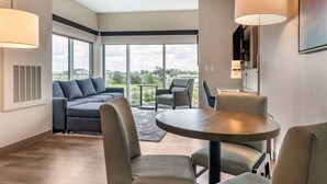 A modern living room with a grey sofa, a round table, and two chairs. Large windows offer a view of the outdoors. - Cambria Hotel Orlando Airport (Orlando)