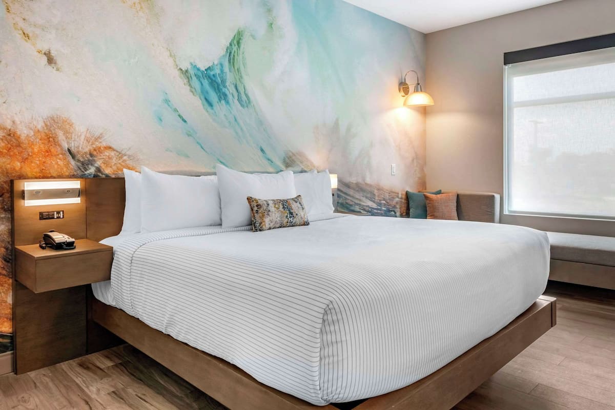 A modern hotel room with a large bed, a nightstand with a phone, a wall-mounted light, and a large window.