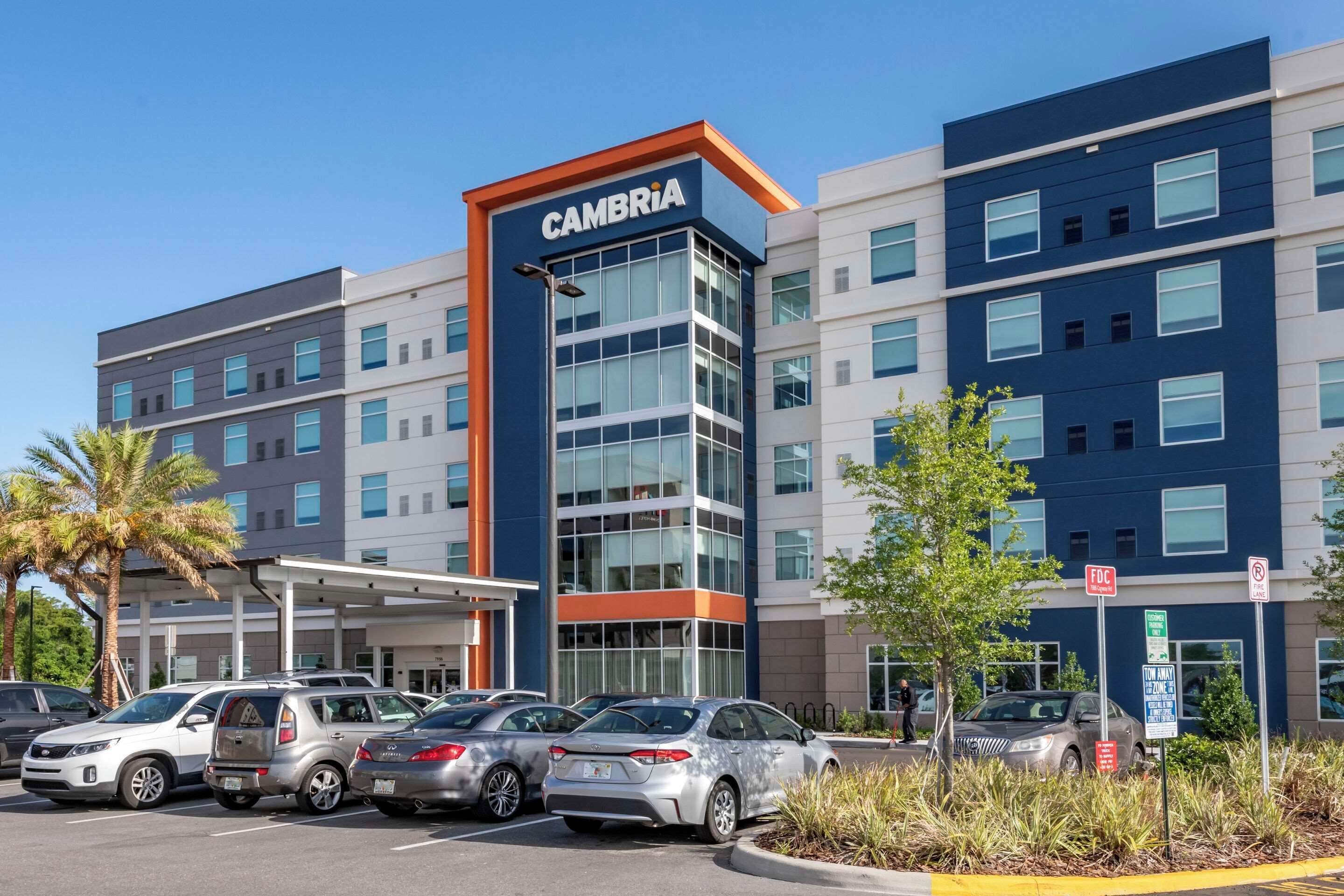 A modern hotel with a prominent CAMBRIA sign, multiple parking spaces, and a clear sky.