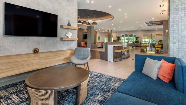 A modern hotel lobby with a large flat-screen TV, a blue sofa, a round wooden table, and a curved reception desk. - Cambria Hotel Orlando Airport (Orlando)
