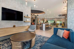 A modern hotel lobby with a large flat-screen TV, a blue sofa, a round wooden table, and a curved reception desk. - Cambria Hotel Orlando Airport (Orlando)