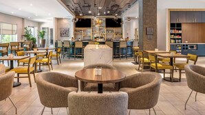 A modern hotel lobby with round tables and comfortable chairs, a bar area with shelves of bottles, and a kitchen area visible to the right. - Cambria Hotel Orlando Airport (Orlando)
