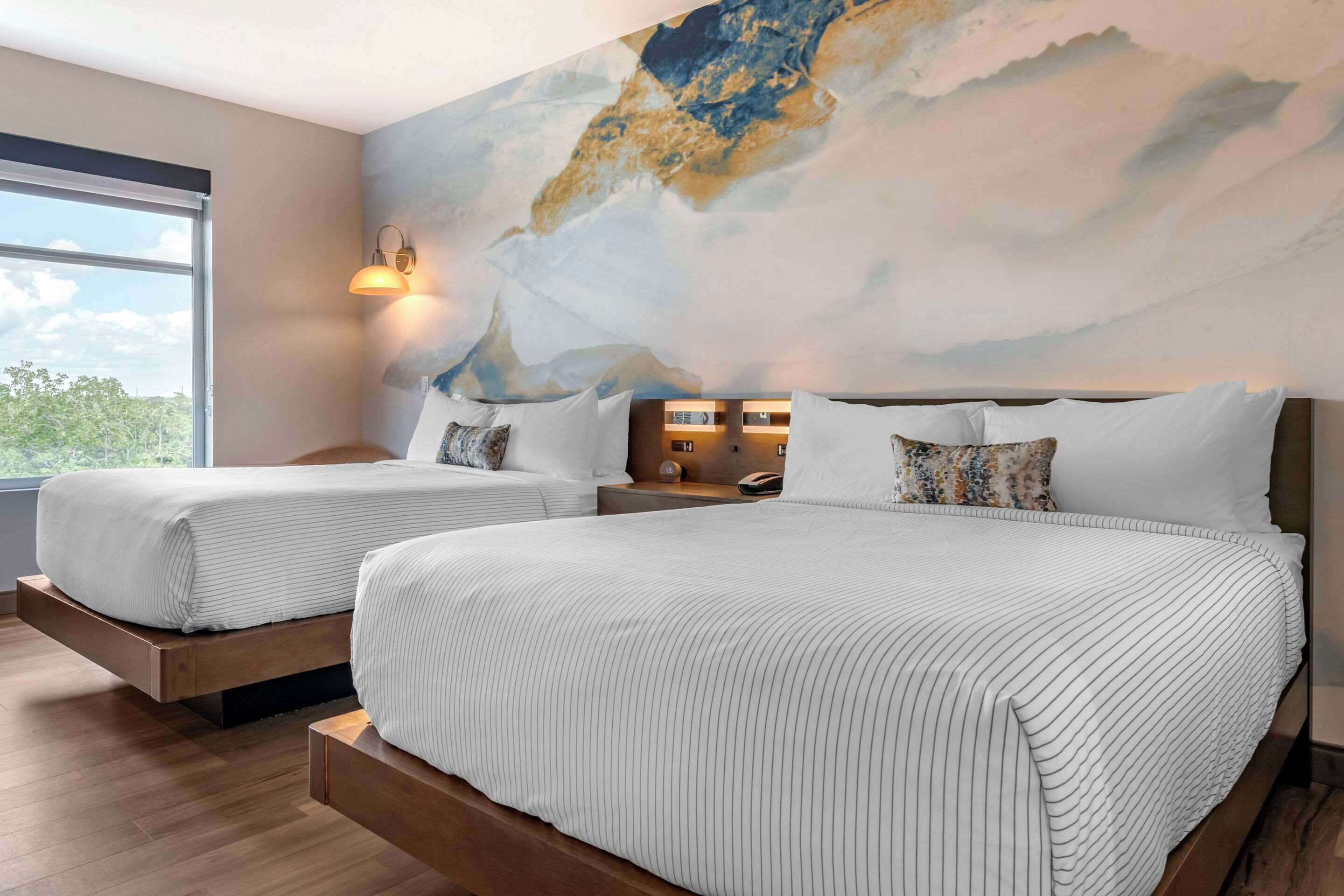 Ein Hotelzimmer mit zwei Betten, einem großen, abstrakten Wandbild, einem Fenster mit Blick auf Bäume und einem Nachttisch mit einer Lampe.