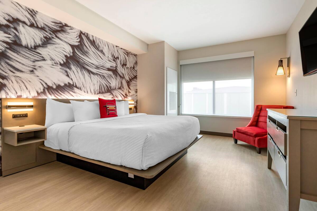 A modern hotel room with a large bed, a red chair, a desk, and a large window.