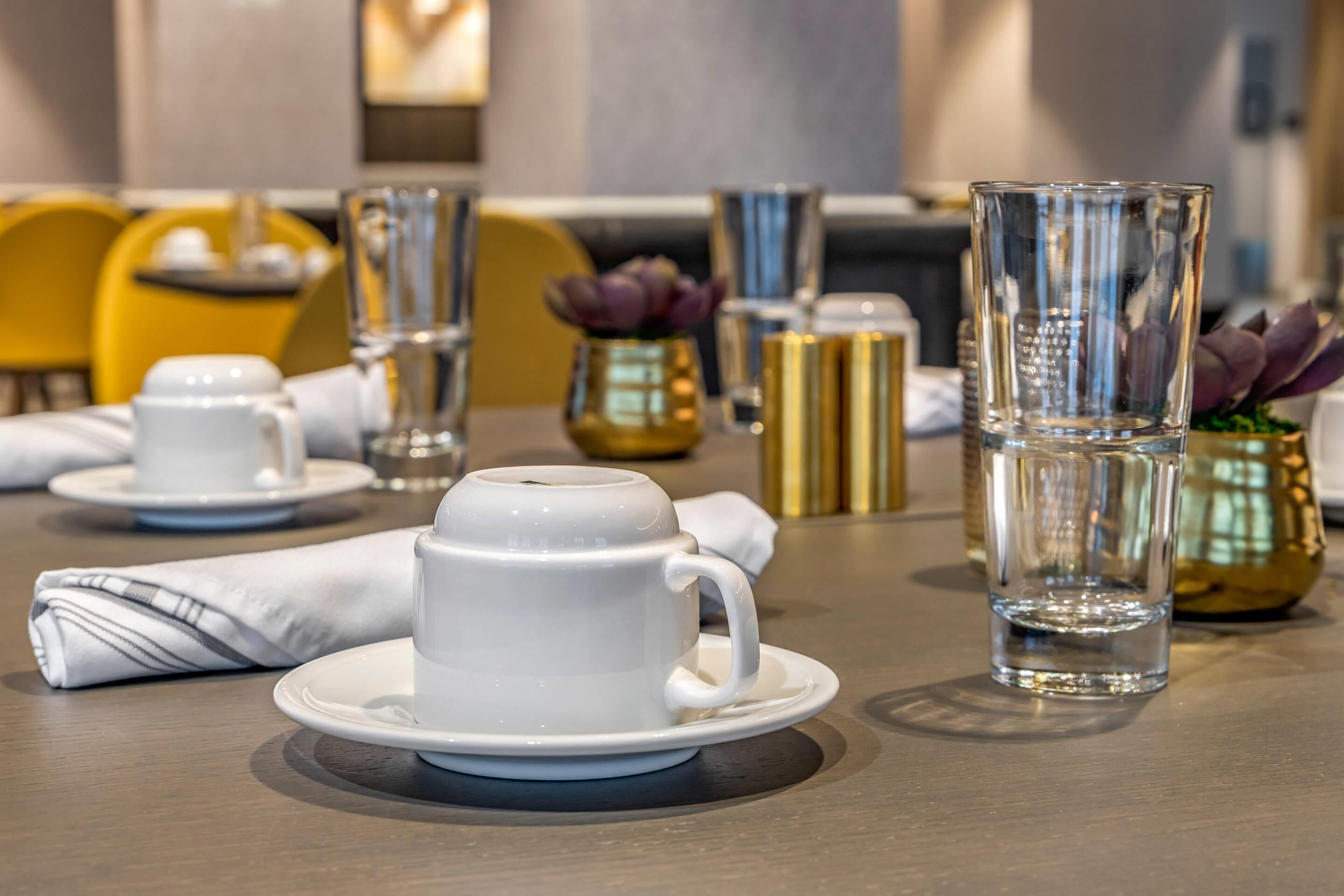A dining table with white cups and saucers, a clear glass, and a decorative vase with purple flowers.