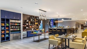 A modern hotel lobby with a self-service food area, seating arrangements, and a bar. - Cambria Hotel Rock Hill - University Center (Rock Hill)