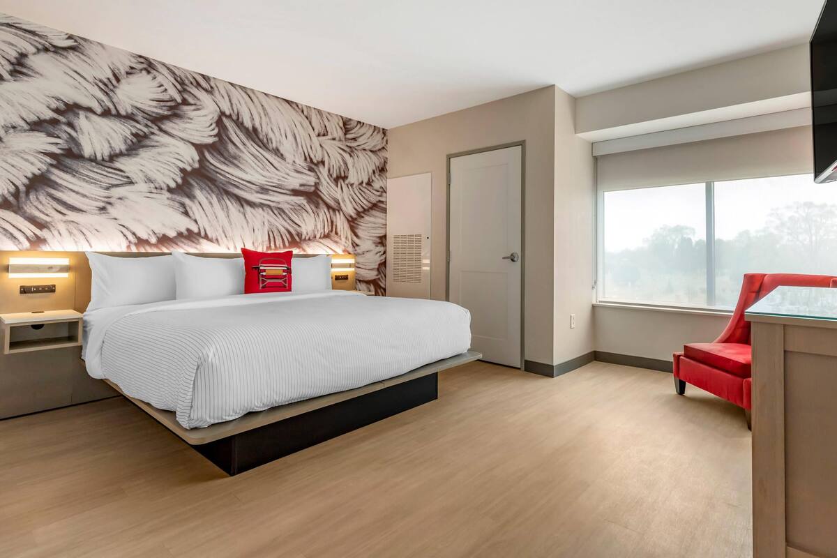 A modern bedroom with a large bed, a red chair, a wall with feather-like wallpaper, and a window with a view.