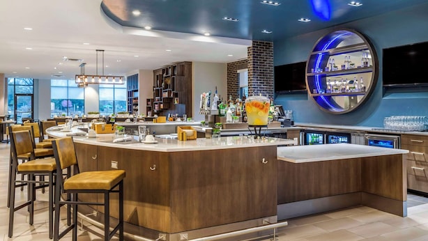 A modern bar area with a curved counter, bar stools, and a large circular mirror with shelves. - Cambria Hotel Rock Hill - University Center (Rock Hill)