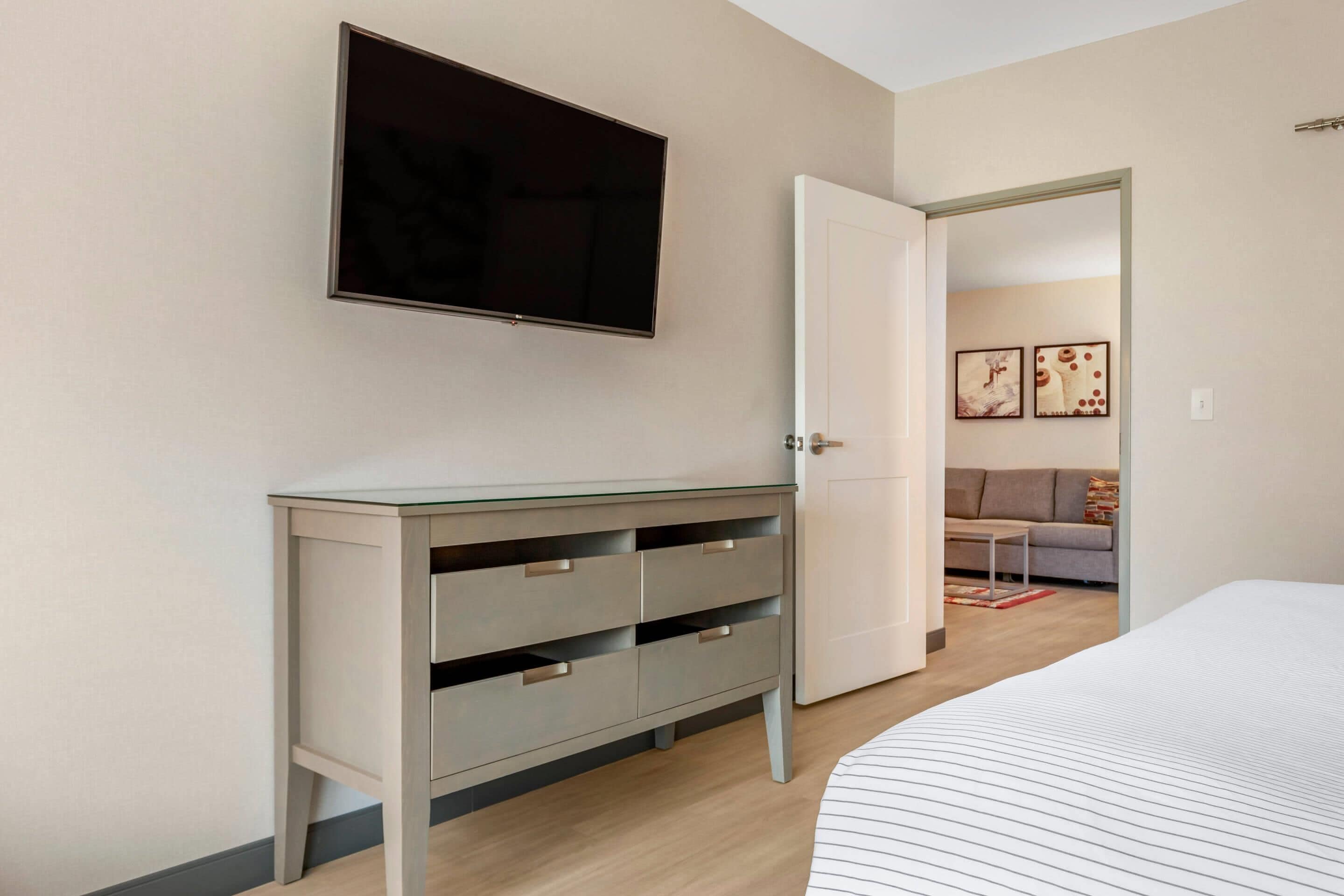 A bedroom with a bed, a TV mounted on the wall, a wardrobe, and a door leading to a living area with a sofa and artwork.