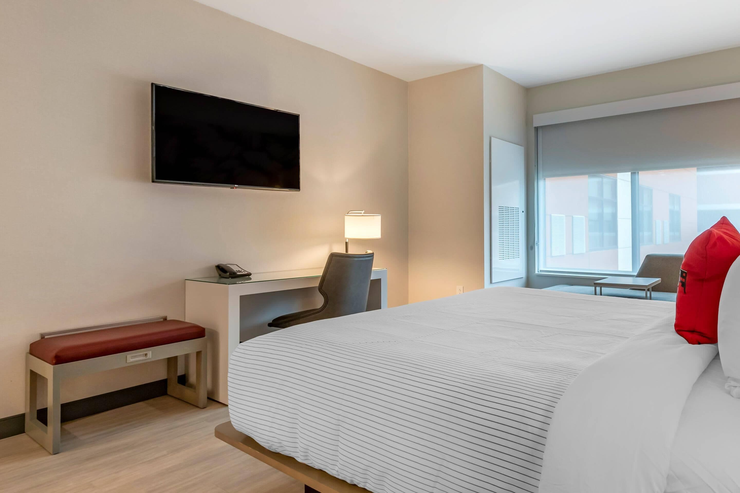 A hotel room with a large bed, a desk with a chair, a TV mounted on the wall, and a window with a city view.