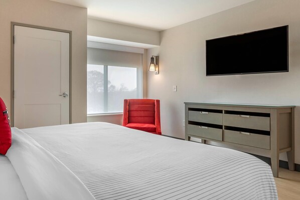 Un dormitorio con una cama, una silla roja, un televisor montado en la pared y un ventanal.