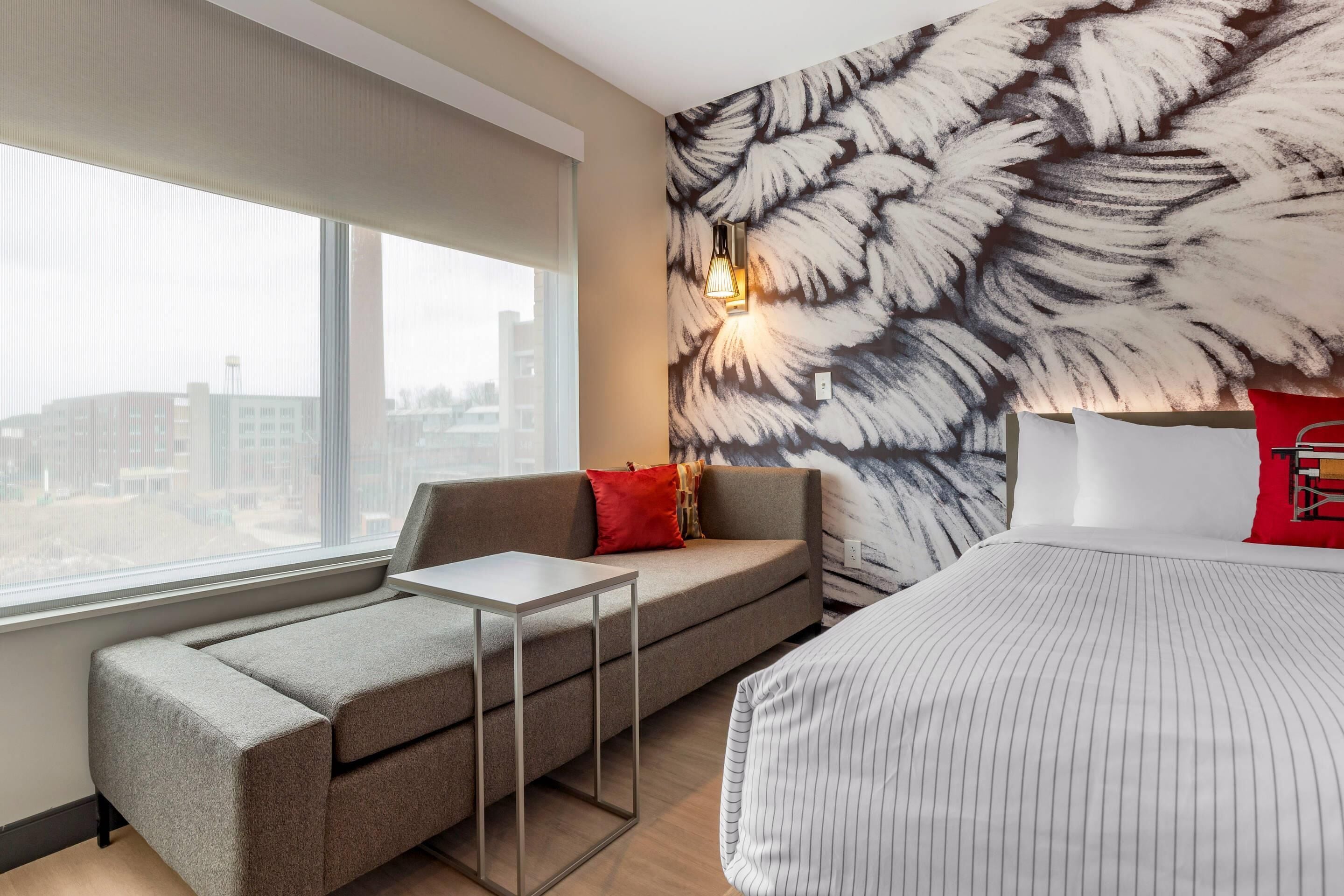 A hotel room with a sofa, a bed, a small table, and a large window.