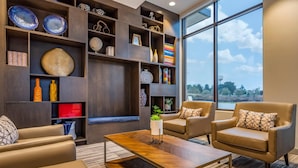 A modern living room with a large window, leather sofas, a wooden coffee table, and a built-in bookshelf with various decorative items. - Cambria Hotel Rock Hill - University Center (Rock Hill)