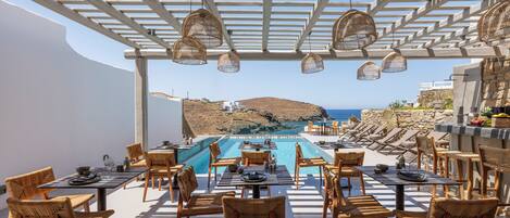 A sunlit outdoor dining area with a pool, wicker chairs, and a view of the sea and hills.