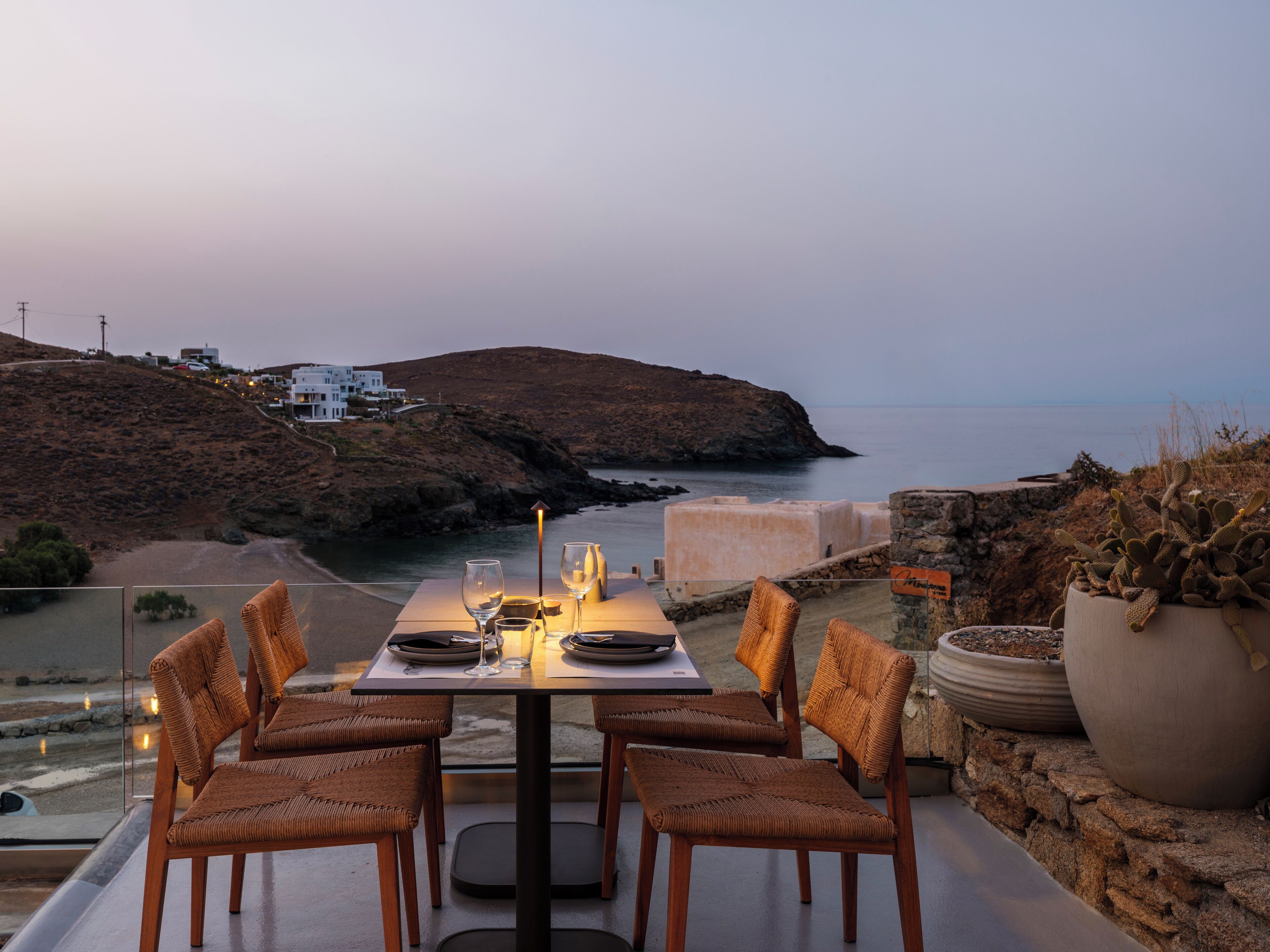 A dining table set with chairs and a candle, overlooking a coastal landscape with buildings and a cliff.