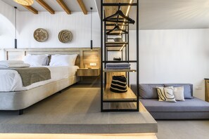 A modern bedroom with a bed, a grey sofa, a wooden shelf with hangers, and a bedside table. - Mykonos Residence Villas & Suites (Mykonos)