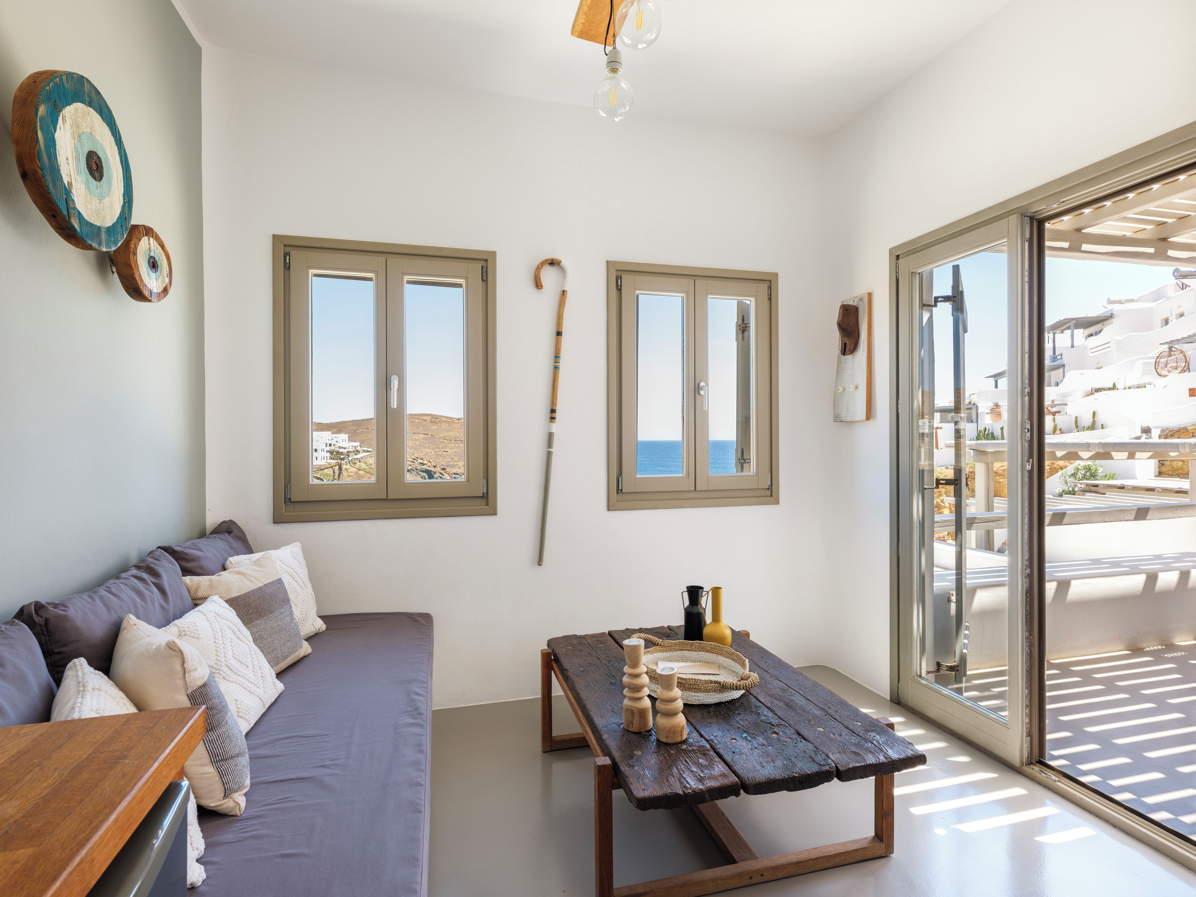 A room with a sofa, a wooden table, and two windows with a view of the sea.