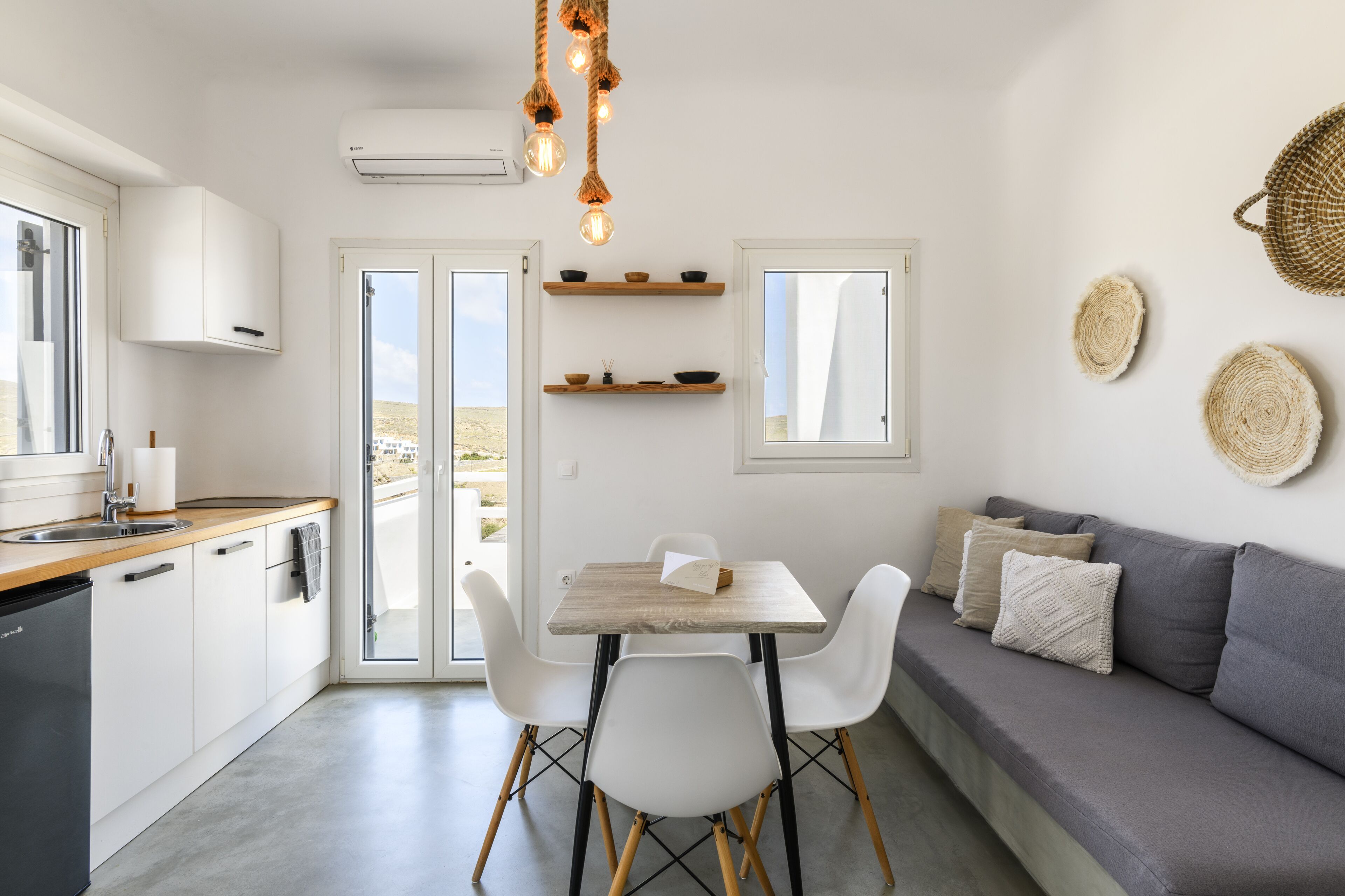 Eine moderne Küche mit weißen Schränken, einer Holzarbeitsplatte und einem Essbereich mit Tisch und Stühlen. Ein graues Sofa mit Kissen und Wandbehänge aus Gewebe sind ebenfalls vorhanden.