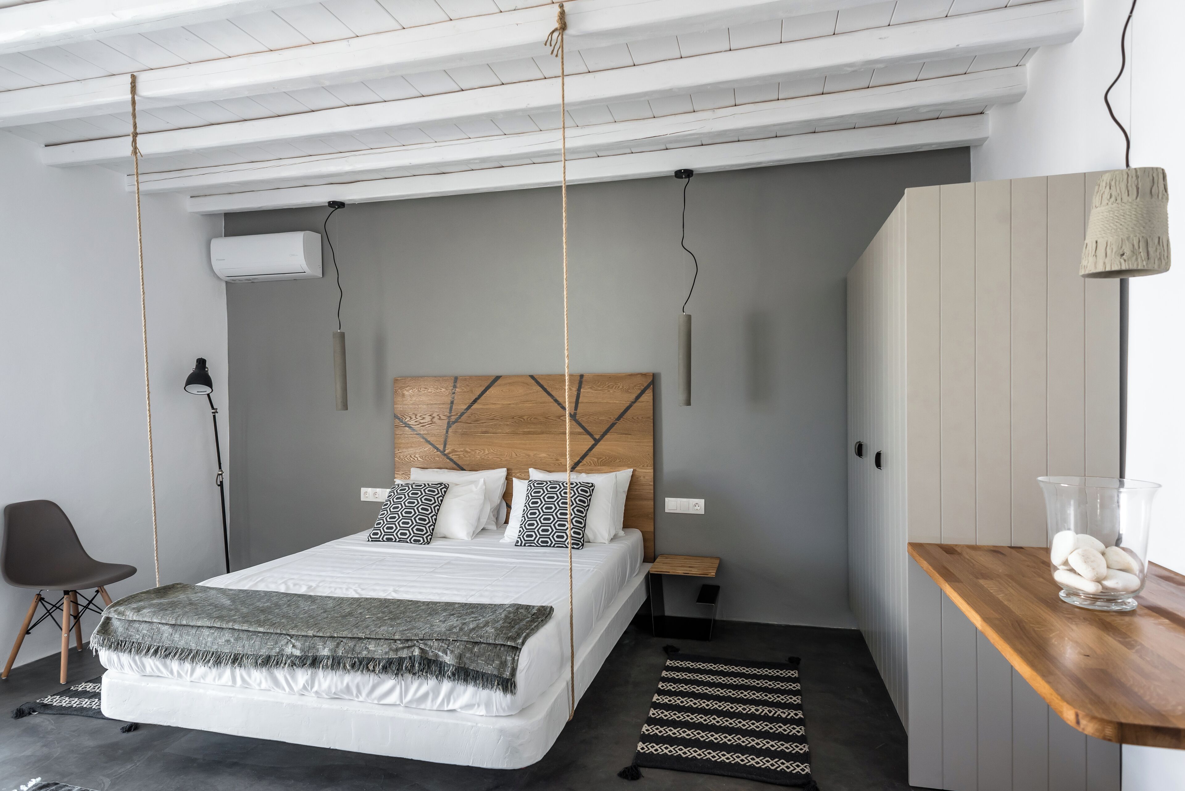 A modern bedroom with a wooden headboard, a bed with patterned pillows, a bedside table, a chair, and a hanging light fixture.