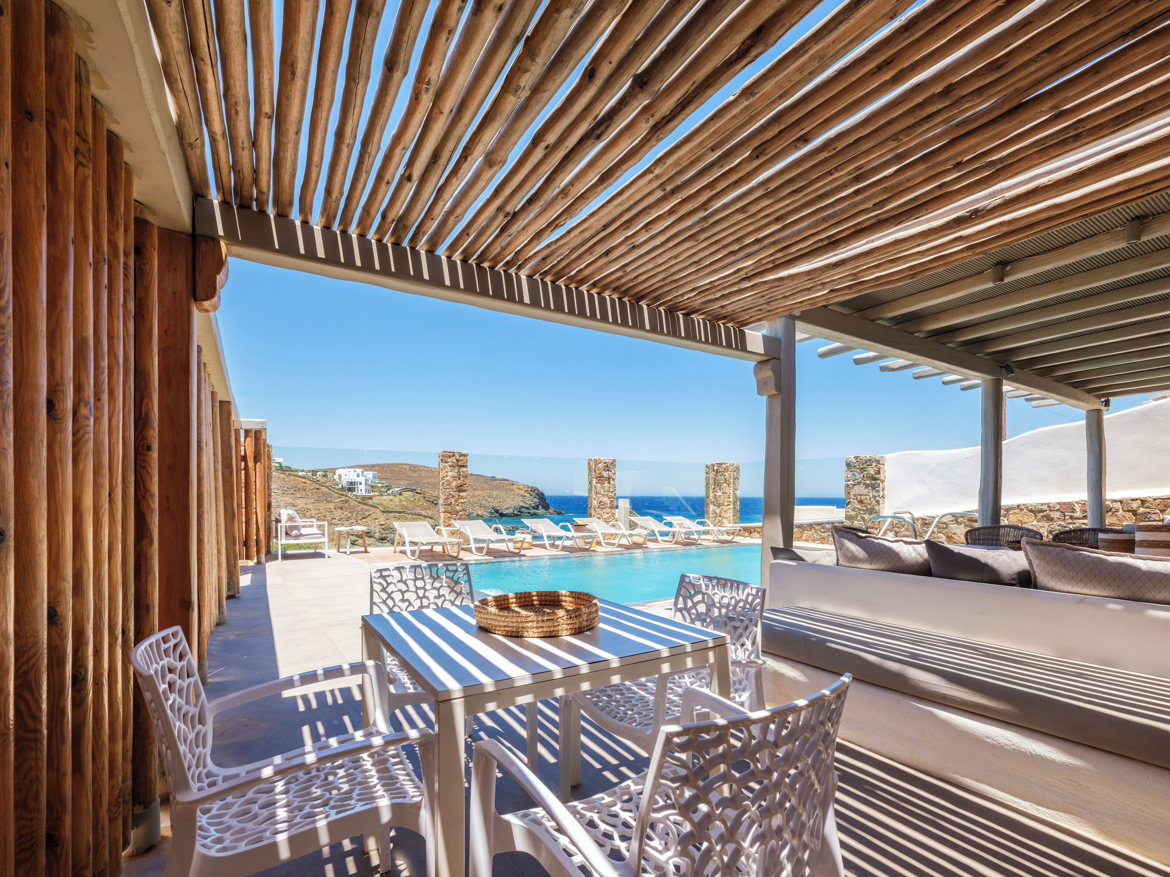 Zona junto a la piscina con sillas de mimbre blanco y una mesa, con vistas al mar azul y un paisaje montañoso.