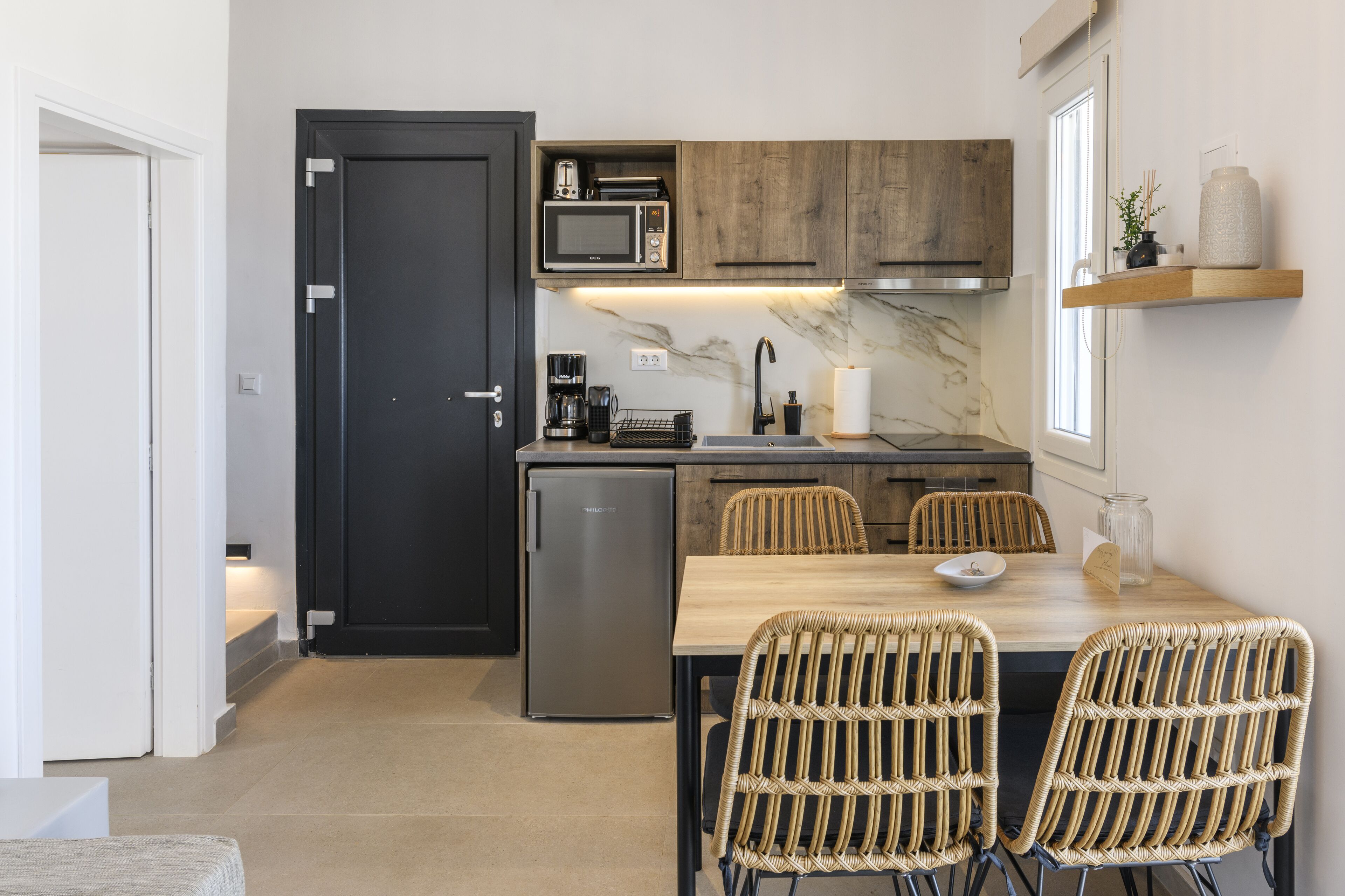 A compact kitchen with a microwave, coffee maker, and a small refrigerator. There is a dining area with wicker chairs and a wooden table.