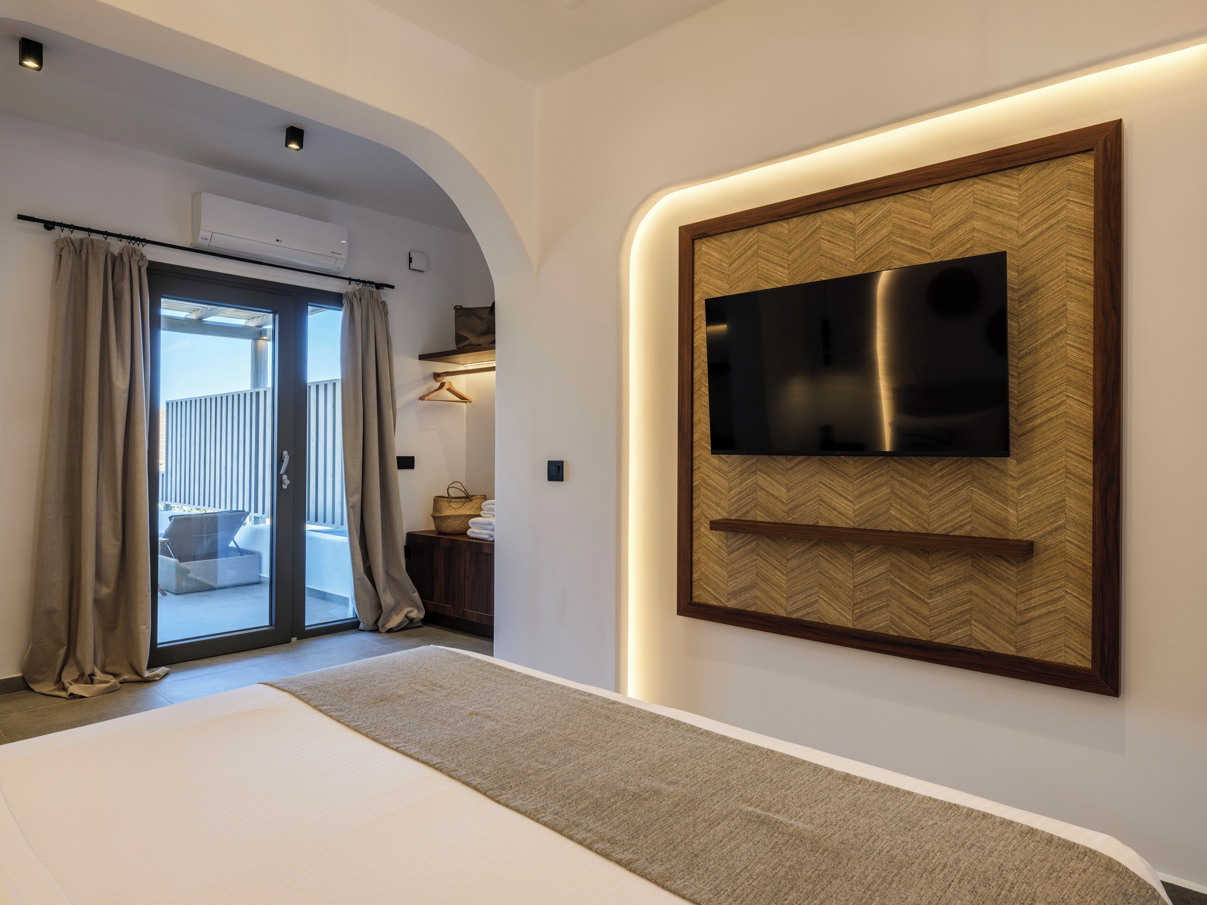 A modern bedroom with a large bed, a TV mounted on a herringbone-patterned wall, and a sliding glass door leading to a balcony.