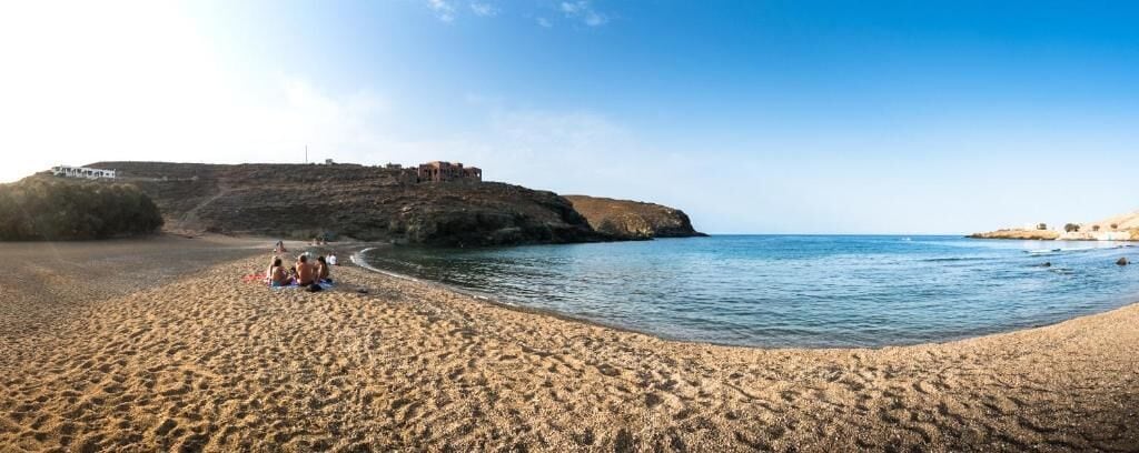 Una playa con gente sentada sobre la arena, un acantilado con edificios y un cielo despejado.