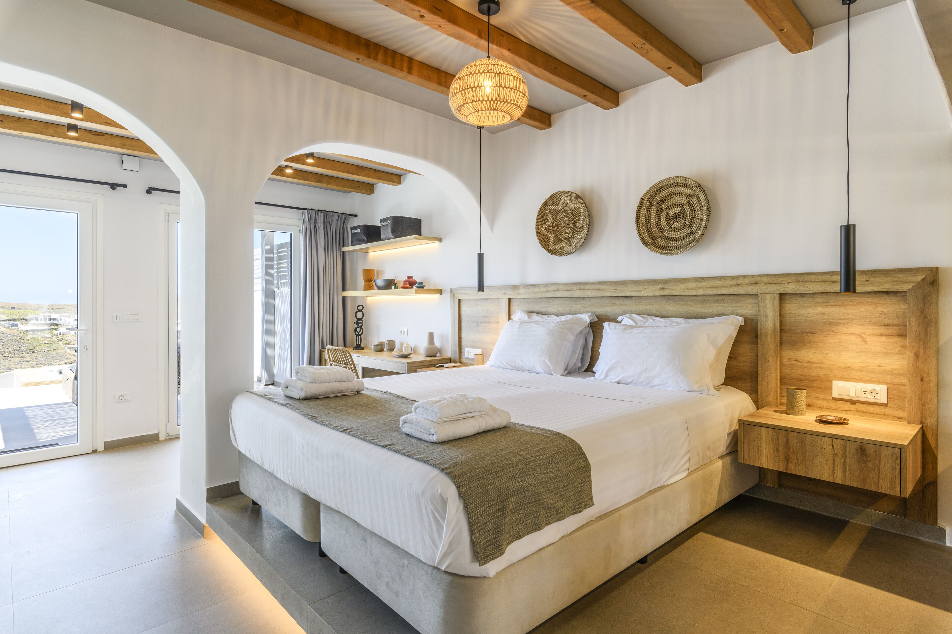 Un dormitorio moderno con una cama grande, cabecera de madera, mesitas de noche, una lámpara colgante y vistas a través de un puerta abierta.