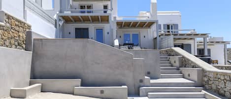 Un edificio moderno con fachada de piedra, una escalera de hormigón que lleva a una terraza de madera y un cielo azul despejado.