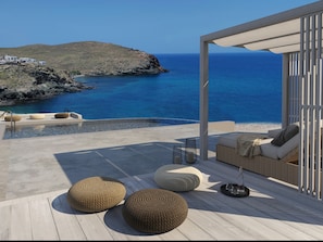 Un área de estar moderna al aire libre con otomanas de mimbre, un sofá y una mesa de vidrio, con vistas a un paisaje costero de aguas azules cristalinas y terreno rocoso.