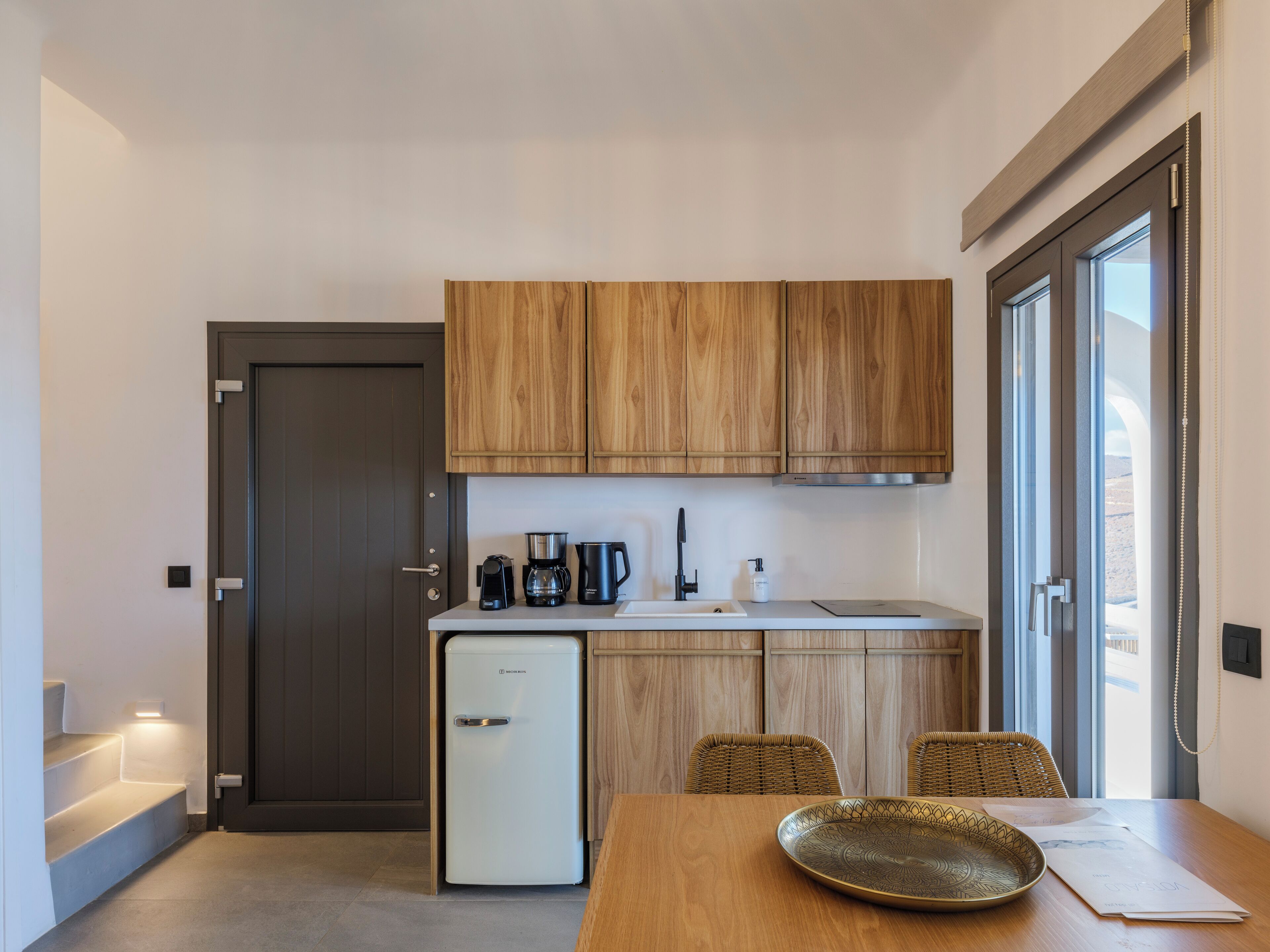 Cocina compacta con armarios de madera, refrigerador empotrado, cafetera y pileta. Hay una mesa de comedor con un plato decorativo y papeles.