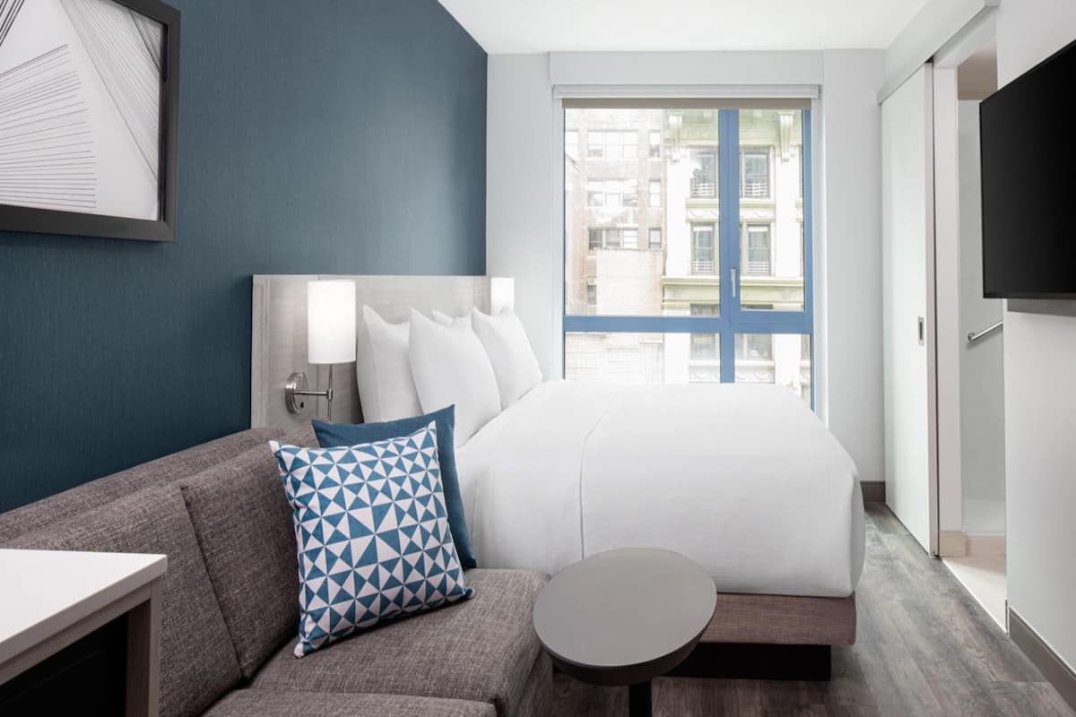 A hotel room with a bed, a sofa, a small table, a TV, and a window with a view of buildings.