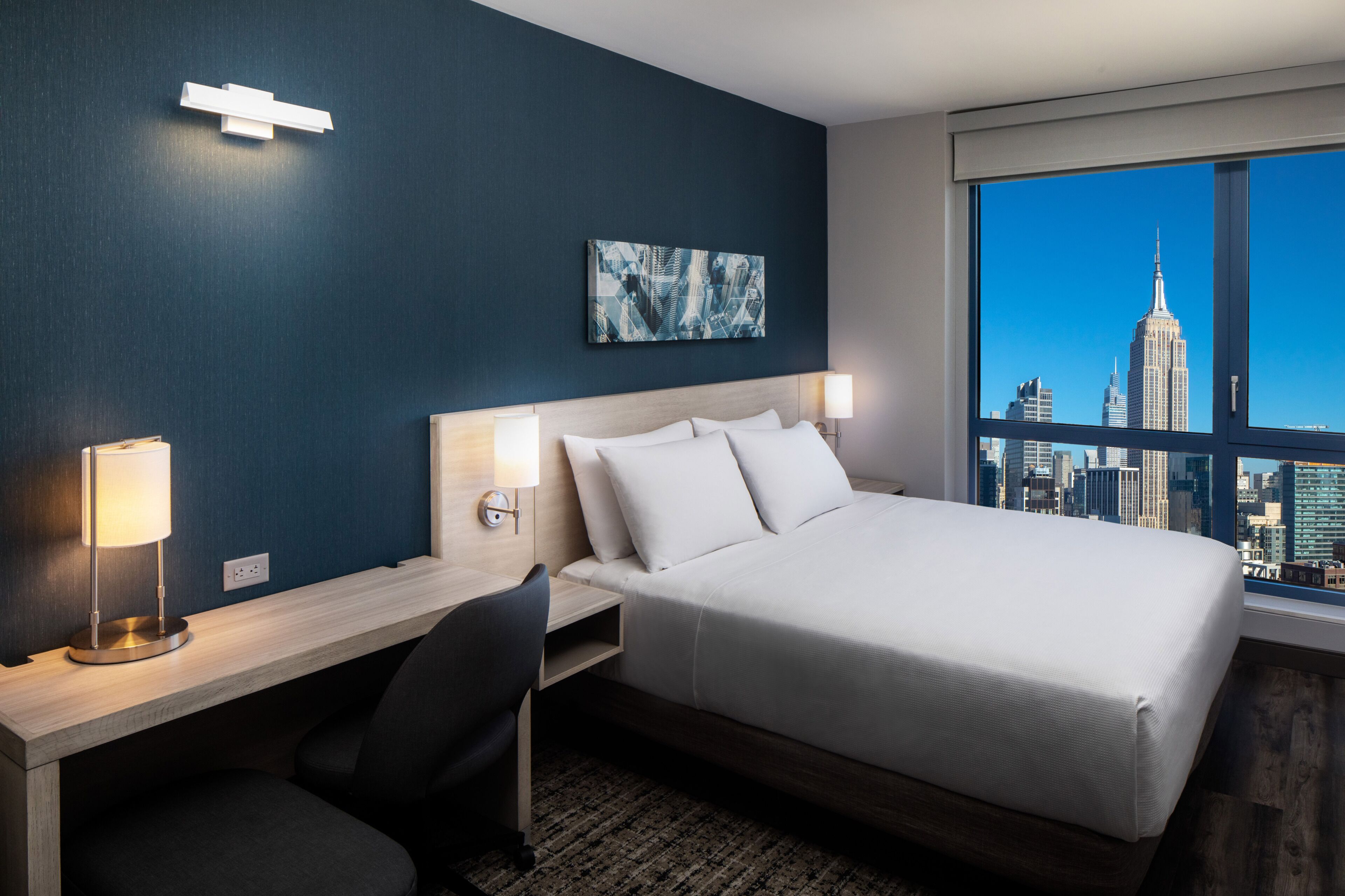 A hotel room with a bed, desk, chair, and a large window with a cityscape view.