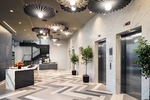 A modern lobby with a reception desk, potted plants, elevators, and unique ceiling lights.