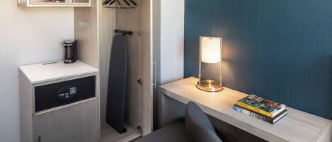 A compact hotel room with a desk, a chair, a coffee maker, a hanging shower curtain, a shelf with a bowl, and a lamp.