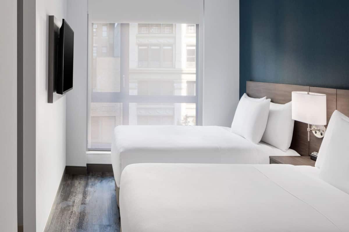 A hotel room with two beds, a TV mounted on the wall, a nightstand with a lamp, and a view of buildings through the window.