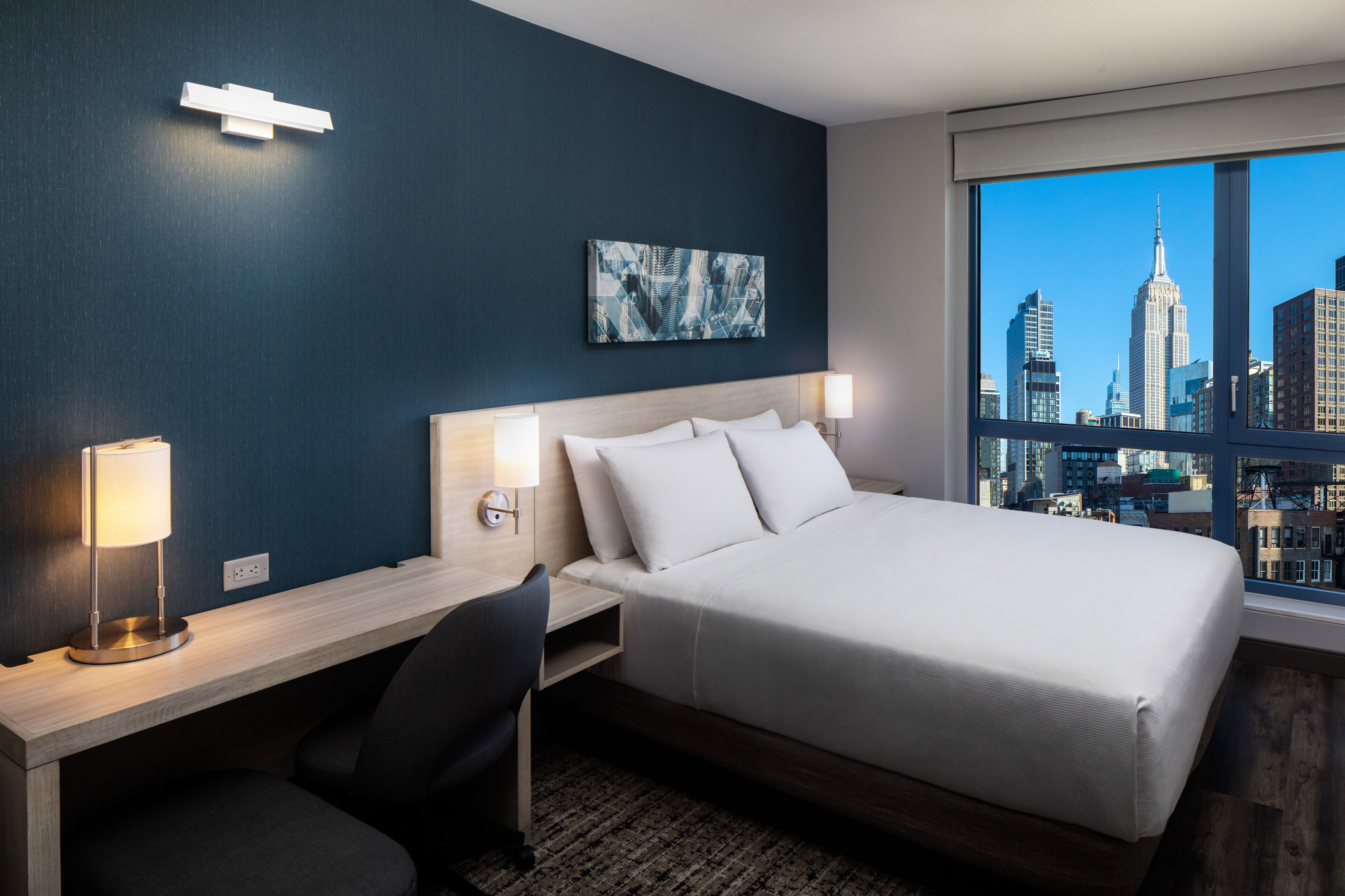 A hotel room with a bed, desk, chair, and a cityscape view through the window.