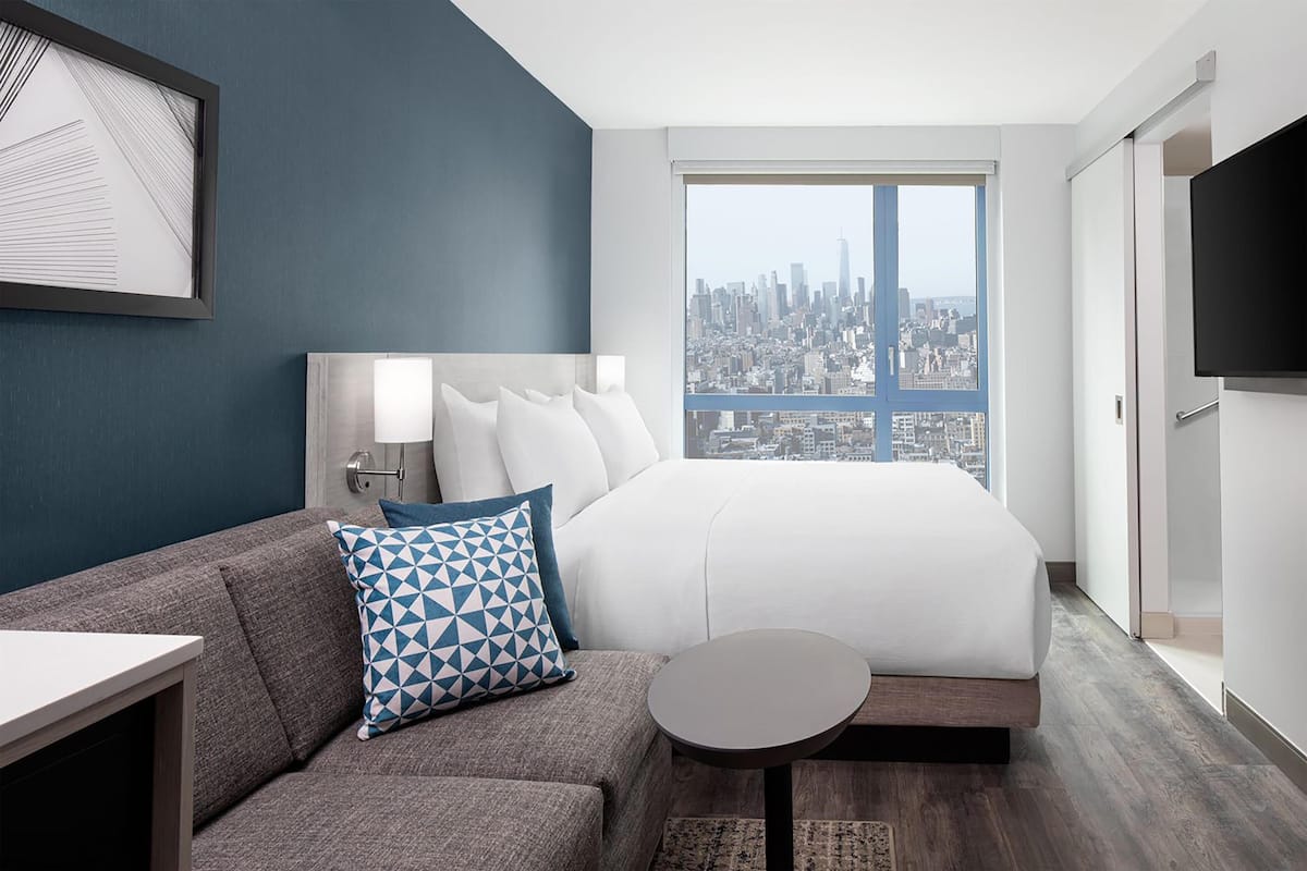 A modern hotel room with a city view, a bed, a sofa, a small table, and a TV.
