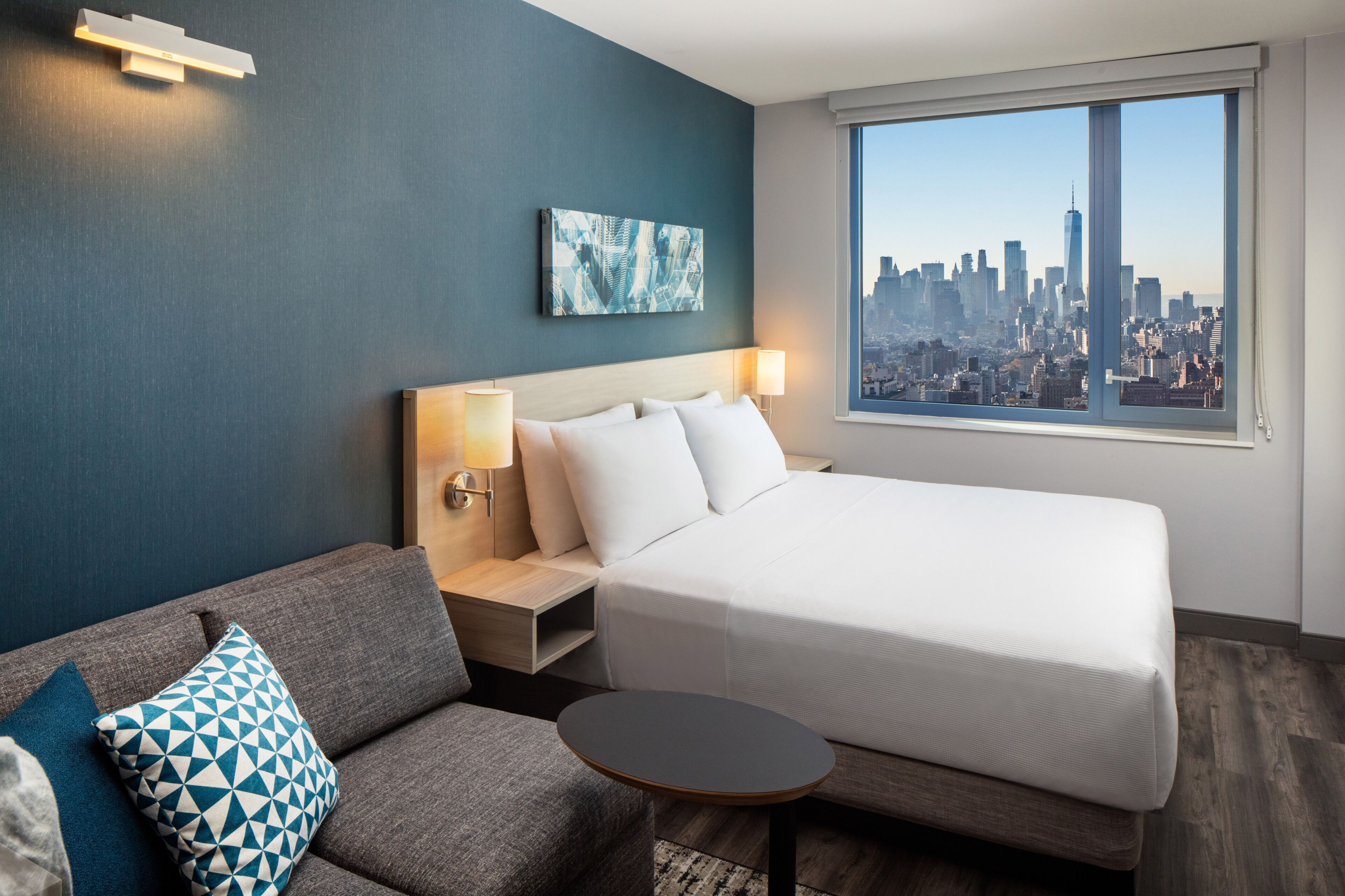 A hotel room with a bed, a sofa, a small table, a nightstand, a lamp, a wall-mounted artwork, and a window with a cityscape view.