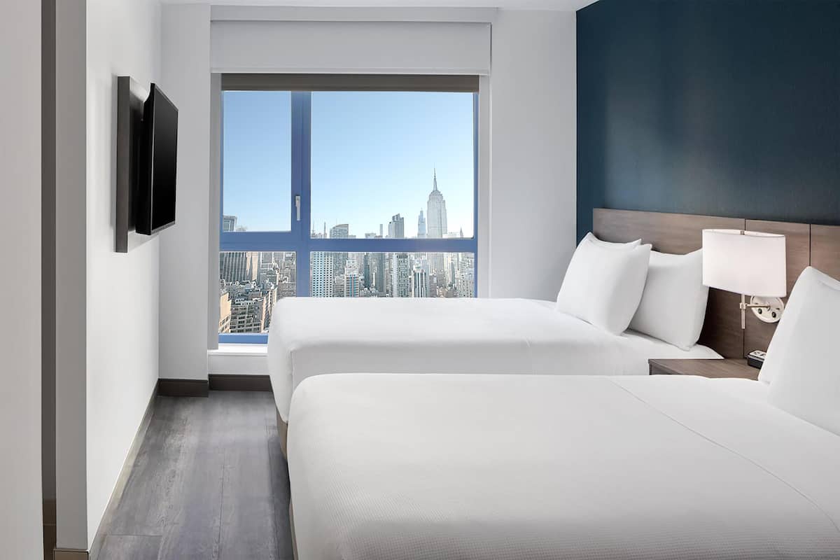 A hotel room with two beds, a TV mounted on the wall, a nightstand with a lamp, and a large window offering a city skyline view.