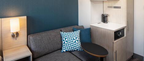 A compact room with a sofa, a small table, a lamp, a coffee maker, and a cabinet.