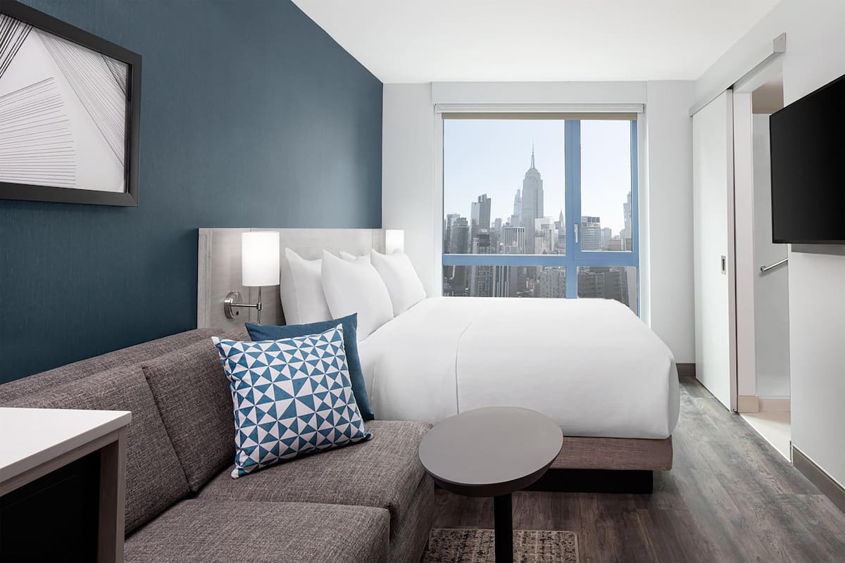 A hotel room with a bed, a sofa, a small table, a TV, and a city view.