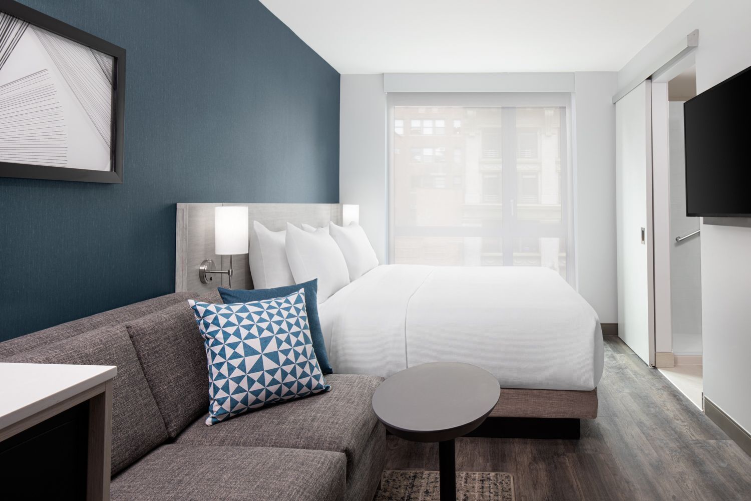 A modern hotel room with a bed, a sofa, a small table, a TV, and a window with a view.