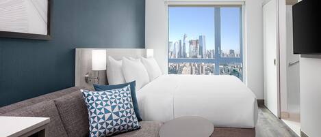 A hotel room with a bed, a sofa, a small table, a TV, and a large window with a city view.