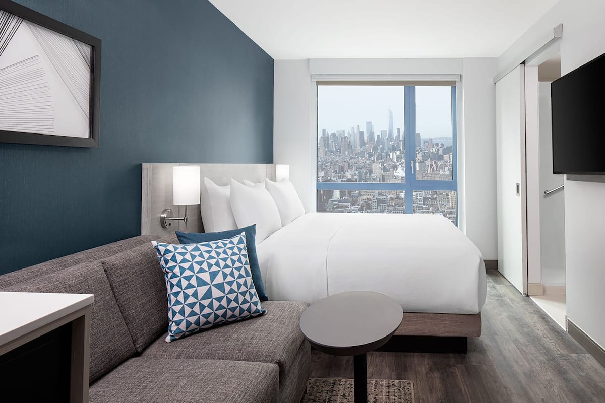 A hotel room with a bed, a sofa, a small table, a TV, and a large window with a city view.