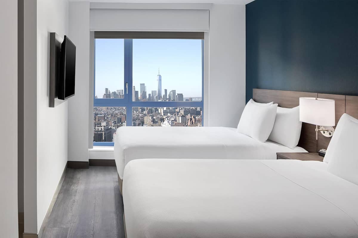 A hotel room with two beds, a TV mounted on the wall, a nightstand with a lamp, and a view of a city skyline.