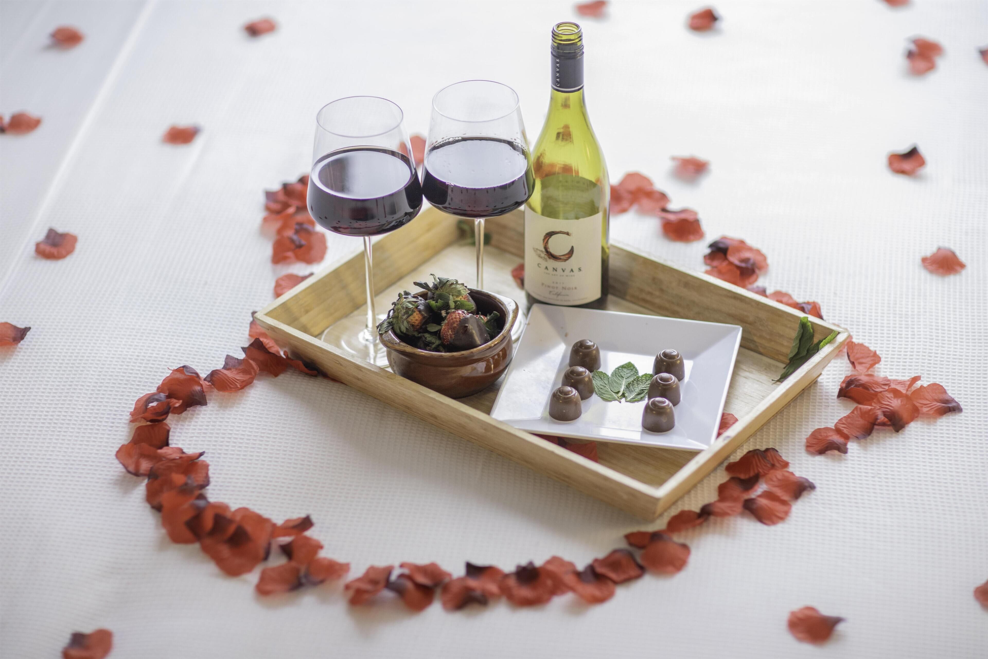 A tray with two glasses of red wine, a bottle of wine, a small bowl with a plant, and a plate with chocolates on a table covered with rose petals.