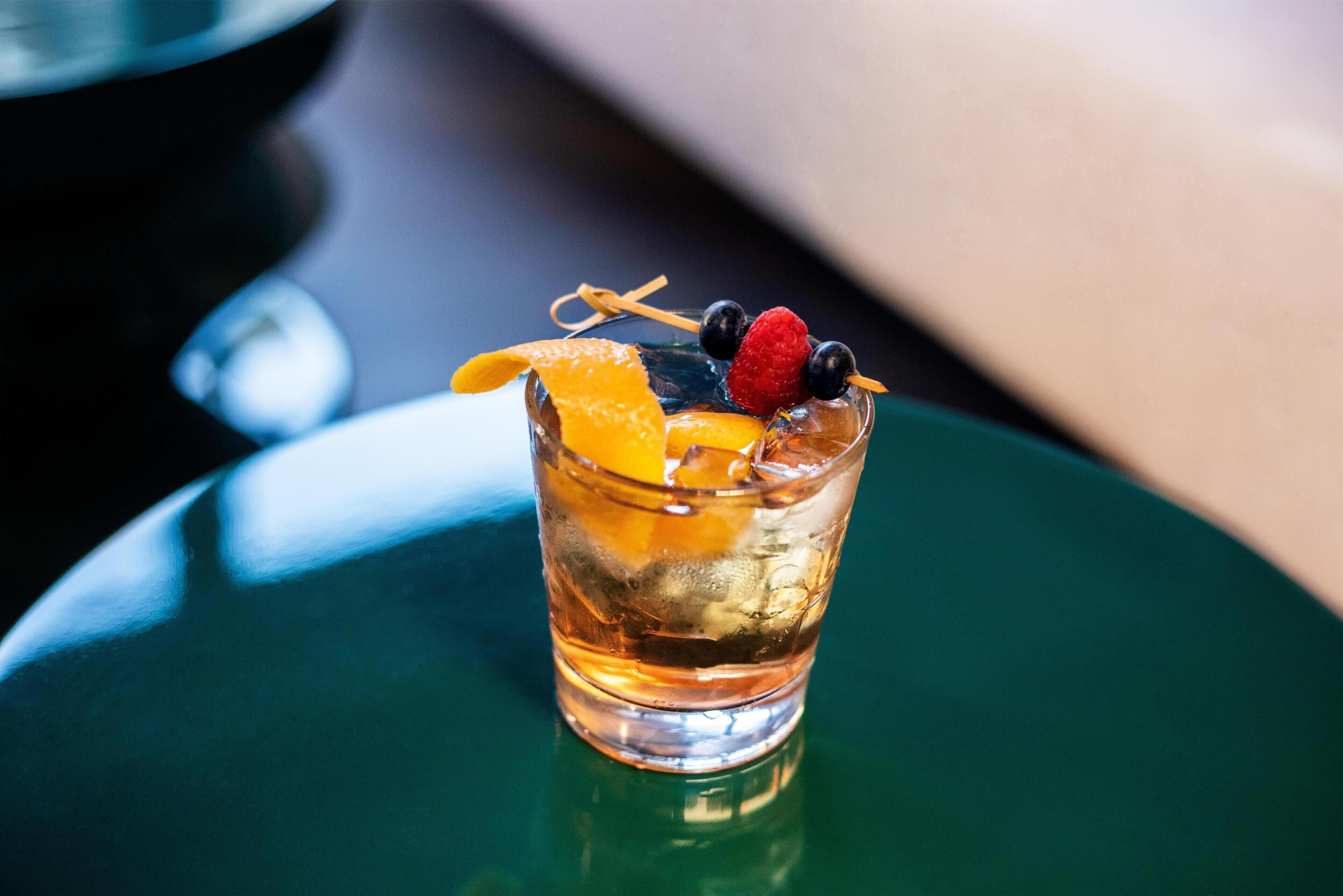 A glass with a beverage, garnished with a slice of orange, a raspberry, and a blueberry, on a reflective surface.