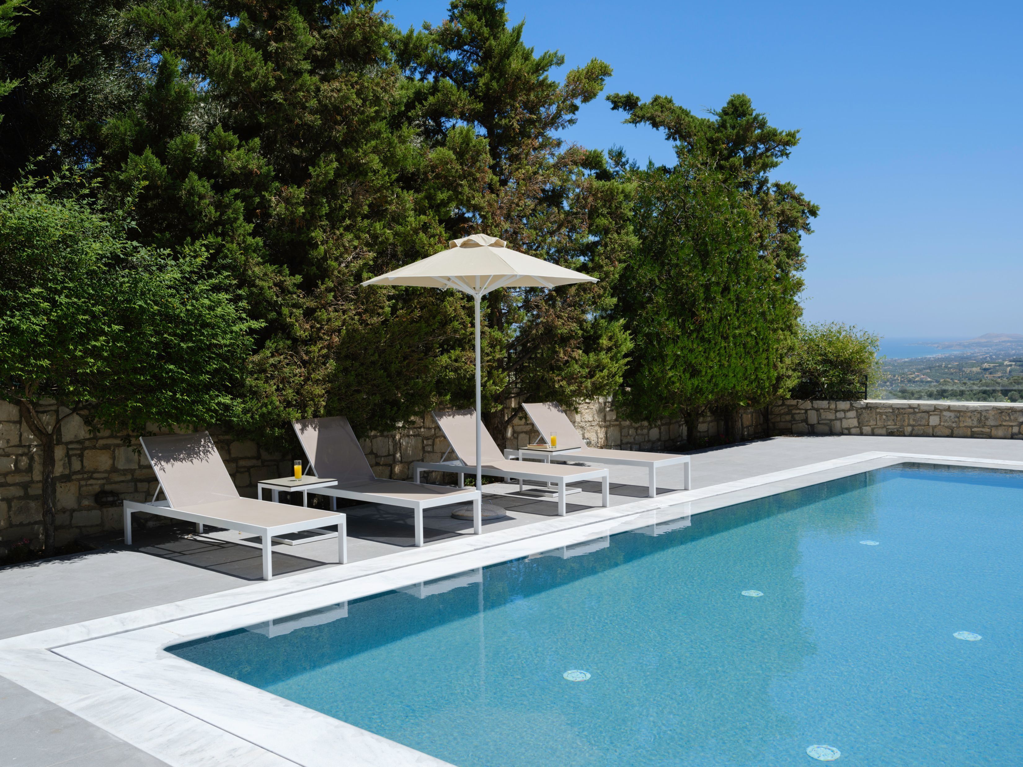 Zona de piscina con sillones de descanso y una mesa, rodeada de muros de piedra y árboles.
